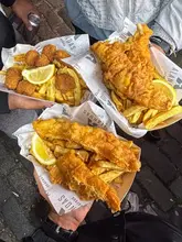 A high-angle, first-person perspective shot showing three paper trays of fish and chips and scampi being held up, likely at a street food market or festival.
