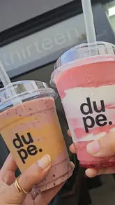 A short, handheld-style video showcasing a visit to 'dupe.', featuring close-ups of a chicken wrap, a protein bowl, protein balls, and smoothies.