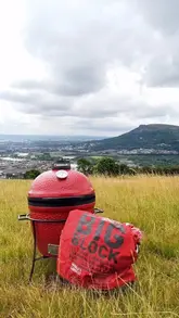 A man prepares a breakfast burrito on a portable Kamado Joe grill while on a hill overlooking Belfast. The video follows the cooking process from lighting the charcoal to assembling the burrito with bacon, sausage, egg, relish, and avocado.