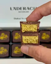 A close-up, macro-style photograph showing a hand holding a halved 'Dubai style' chocolate praline, clearly displaying the pistachio kunafa filling, with the branded Läderach box visible in the background.