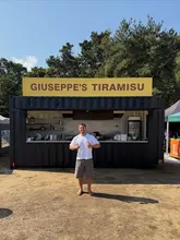 A man stands in front of a black shipping container food stall branded 'Giuseppe's Tiramisu' at an outdoor festival, giving a thumbs up to the camera.