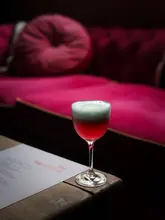 A moody, atmospheric shot of a cocktail in a coupe glass, set against a backdrop of pink velvet seating at The Blackbird in Edinburgh. The lighting is low and intimate, highlighting the drink's foam while keeping the venue's interior soft and out of focus.