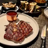 A high-quality, moody, and professional shot of a premium steak dinner at a restaurant, featuring a well-seared ribeye, roasted bone marrow, and accompanying sides, effectively capturing the essence of a luxury dining experience.