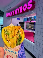A close-up, handheld shot of a gyro held in front of the Lucky Gyros stall in the Manchester Arndale food court, showcasing the branding and the product.