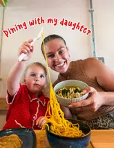 A candid, family-focused shot of a parent and child dining at a casual street food venue, featuring a playful noodle pull and gyoza.
