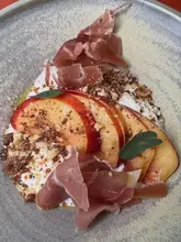 A high-quality, overhead food shot featuring a fresh dish of sliced peaches, prosciutto, and a creamy base, garnished with a crumble topping. The image is bright, clean, and professionally styled, effectively showcasing the freshness of the ingredients.