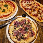 An overhead flat-lay photograph of three different pizzas served on a wooden table, with one pizza specifically labeled as gluten-free.