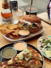 A high-quality, well-lit food photography shot featuring a Mexican-inspired meal including a brisket-style sandwich, tortilla chips with dip, and tacos, accompanied by a bottle of Modelo beer.