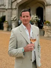 A portrait of a man in a suit holding a glass of rosé wine, standing in front of a large stone manor house, likely at a wedding or upscale event.
