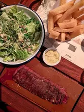 A top-down, overhead shot of a steak meal at Flat Iron, featuring a sliced flat iron steak served on a wooden board, a bowl of green salad, a small pot of sauce, and a metal cup of chips.