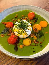 A vibrant, overhead shot of a burrata dish served in a green gazpacho, garnished with barbecued tomatoes, hazelnuts, and curry leaves. The lighting is warm and even, highlighting the textures of the cheese and the freshness of the sauce.