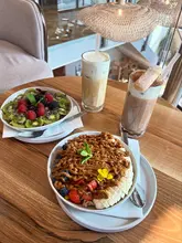 A high-quality, overhead flat-lay photograph of a brunch spread featuring two acai bowls—one topped with fresh berries and one with lotus biscoff—alongside two iced lattes, set on a wooden table in a cafe setting.