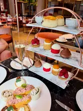 A high-quality, professional promotional shot of an afternoon tea service at a luxury hotel. The image effectively captures the tiered stand and a plate of savoury items, using a shallow depth of field to draw focus to the food while maintaining a clean, elegant background.
