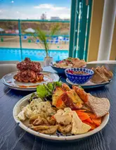 A vibrant, well-lit shot of a lunch spread at a lido restaurant, featuring a variety of dishes including chicken and waffles, shakshuka, and a mezze platter, with a scenic pool and sea view in the background.