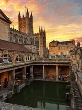 A picturesque, high-quality shot of the Roman Baths in Bath, England, taken during a golden hour sunset. The composition effectively frames the historic pool and the Bath Abbey in the background, creating a serene and inviting atmosphere for visitors.