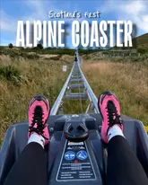 A first-person perspective shot of an alpine coaster track in the Pentland Hills, Scotland, featuring the rider's legs and feet in the foreground. The image effectively captures the sense of adventure and the scenic outdoor setting, with clear text overlay identifying the attraction.