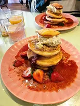 A high-quality, appetizing shot of strawberry shortcake pancakes at a brunch venue, featuring a stack of pancakes with coulis, chantilly cream, and a madeleine, accompanied by drinks.