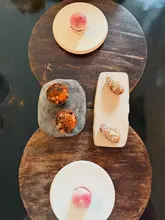 A high-quality, top-down flat-lay photograph showcasing a selection of refined small plates from a tasting menu, arranged symmetrically on wooden boards. The image highlights the intricate presentation of the dishes, including macarons and pastries, against a dark, clean background.