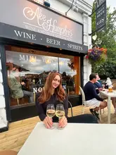 A lifestyle shot featuring a woman enjoying a glass of white wine on the outdoor terrace of Ake & Humphris, a wine and beer specialist venue. The image captures a relaxed, social atmosphere suitable for casual drinks.