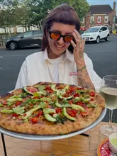A casual, influencer-style shot featuring a person holding a pizza in front of their face while dining outdoors at a pub. The image captures a relaxed, social atmosphere with a focus on the unique pizza toppings.