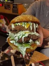 A close-up, eye-level shot of a double-patty fried chicken burger being held by a person, featuring a generous amount of sauce and lettuce.