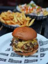 A close-up, 45-degree angle shot of a smash burger served on branded paper, with a basket of fries in the background. The image captures the messy, indulgent nature of a classic smash burger, focusing on the melted cheese and caramelized onions.