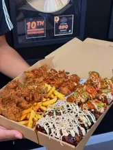A first-person, eye-level shot of a large, overflowing 'Feast Box' containing various styles of chicken wings, tenders, and loaded fries, held up against a wall displaying award certificates.
