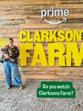 A lifestyle photo featuring a creator posing with their dog in front of the Diddly Squat Farm Shop sign, associated with the TV show Clarkson's Farm. The image captures a casual, authentic moment during a road trip, highlighting the venue as a tourist destination.