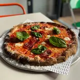 A high-quality, well-lit photograph of a freshly baked pizza, likely a pepperoni or salami style, served on a metal tray with parchment paper. The image captures the texture of the crust and the melted cheese, presenting the food in an appealing, casual dining setting.