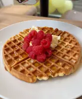 A close-up, top-down shot of a waffle topped with fresh raspberries and cream, with syrup being poured over it. The setting appears to be a casual cafe environment.