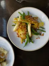 A high-quality, overhead shot of a vibrant peach and green bean salad, garnished with a spiced almond crumb, served on a white plate against a dark, minimalist table surface.