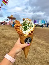 A handheld, close-up shot of a fried chicken and waffle cone topped with sauce and chives, held against a blurred festival background featuring a carousel.