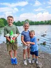 A bright, family-focused photograph taken at a park by a lake, capturing a day out with children.