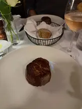 A high-quality, focused shot of a fillet steak served on a white plate, captured in a restaurant setting. The background features a glass of wine and a bread basket, creating a warm, atmospheric dining scene.