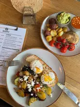 A top-down, overhead shot of two brunch plates and a menu on a wooden table at Occombe Farm. The dishes include a vegetarian breakfast with poached eggs and a plant-based breakfast with beetroot falafel.