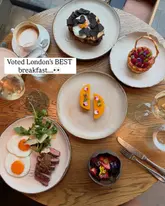 A high-quality, overhead flat lay photograph of an upscale breakfast spread at HIDE restaurant in London. The image features a variety of beautifully plated dishes, including steak and eggs, a delicate pastry, and fresh fruit, arranged on a wooden table with cutlery and drinks, conveying a sophisticated and premium dining experience.