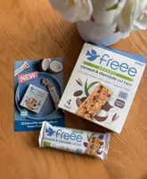A flat-lay product shot featuring a box of Freee organic coconut and chocolate oat bars, a single bar, and a promotional flyer on a wooden table, accented by white flowers.
