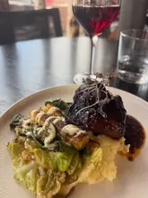 A close-up, slightly high-angle shot of a BBQ lamb shank dish served with greens and a side of sauce, accompanied by a glass of red wine. The lighting is functional but lacks professional polish, and the composition feels somewhat cramped due to the cropping of the plate and glass.