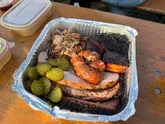 A top-down shot of a BBQ meat platter served in a foil takeaway container on a wooden table, featuring pork belly, brisket, turkey, sausage, and pickles.