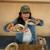 A casual, selfie-style photo of a creator at a wine bar, holding up plates of oysters to demonstrate their size, with a side of pickled string beans visible on the table.