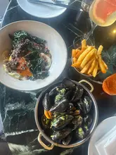 A high-quality, overhead flat-lay shot of a meal at Fallow in Soho, featuring flamed mussels, kombu-cured bass, and fries, accompanied by a cocktail. The lighting is natural and flattering, highlighting the textures of the food against a dark marble table.