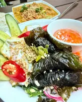 A top-down, natural-light shot of a Vietnamese rice bowl dish featuring grilled meat wrapped in leaves, a fried egg, fresh cucumber, and chili, accompanied by a bowl of curry in the background. The image captures the vibrant colors and textures of the meal, emphasizing the freshness of the ingredients.
