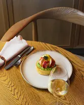 A beautifully composed, high-quality photograph of a lobster benedict dish served on a minimalist plate, set against a clean wooden table and chair. The lighting is soft and natural, highlighting the textures of the food, while the shallow depth of field keeps the focus squarely on the dish, creating an elegant and inviting atmosphere.