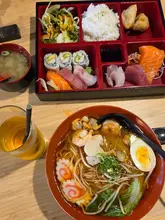 A top-down, flat-lay style photograph of a Japanese lunch spread featuring a bowl of seafood ramen and a sashimi bento box, accompanied by miso soup and a drink on a wooden table.