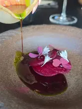 A high-quality, close-up macro shot of a fine dining dish being finished with a sauce pour. The composition focuses on the interaction between the sauce and the plated beetroot and floral garnish, highlighting the artistic presentation and attention to detail.