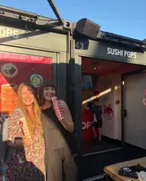 A photo of two people holding branded sushi packaging in front of a 'Sushi Pops' container unit at Boxpark Shoreditch.
