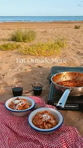 A short, aesthetic video showing two people cooking pasta on a portable stove on a beach, enjoying wine, and watching the sunset with their dog.