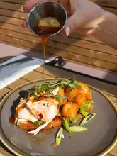 A high-quality, well-lit action shot of a brunch dish being drizzled with sauce. The composition is effective, capturing the texture of the food and the motion of the pour, making it highly appealing.
