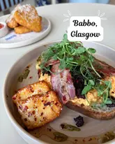 A close-up, top-down shot of a brunch dish featuring scrambled eggs, bacon, black pudding, and crispy halloumi on sourdough, with a side of hash browns, taken at Babbo in Glasgow.