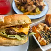 A close-up, handheld shot of a cheeseburger, with a plate of loaded fries and a red drink visible in the background, used to promote a burger deal and drink offer at a restaurant.
